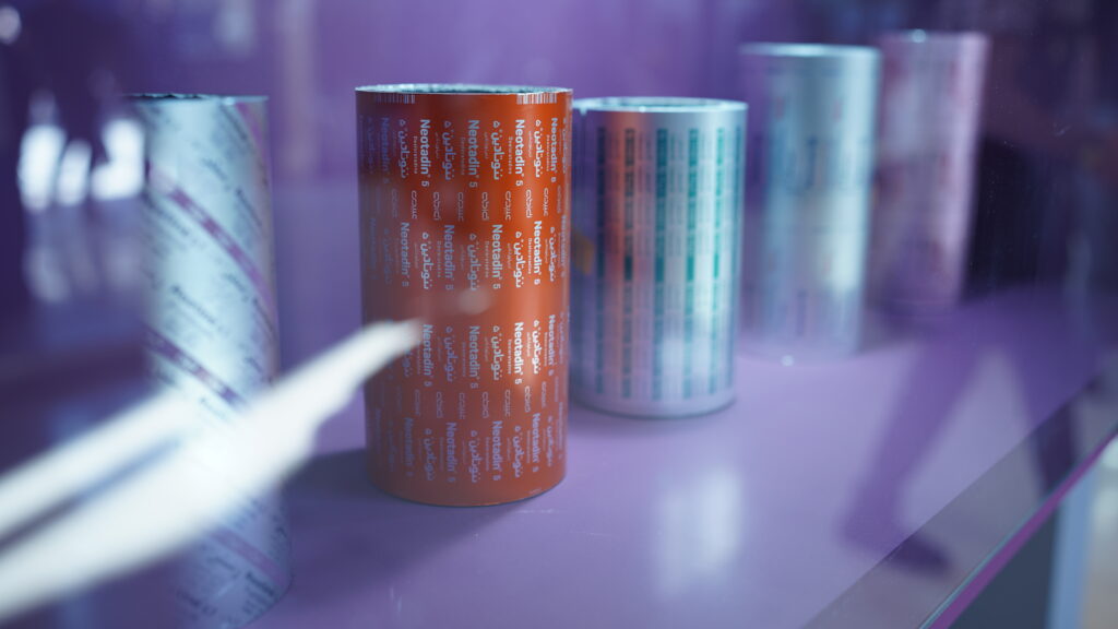 "رول‌های فویل آلومینیومی چاپ‌شده با برند نئوتادین، مناسب برای بسته‌بندی دارویی، نمایش داده‌شده در یک ویترین با پس‌زمینه بنفش."